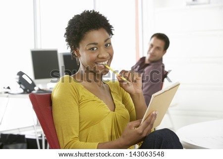 Portrait of smiling young businesswoman holding pencil and notepad with colleague in background