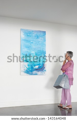 Full length of senior woman with shopping bags observing painting in art gallery