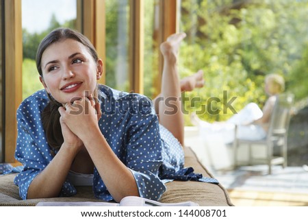 Young woman with hands clasped to chin lying by windows overlooking forest