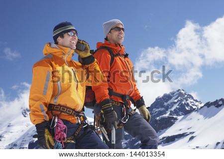 Two male mountain climbers on snowy peak against sky with one using walkie talkie