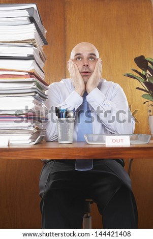 Overworked businessman sitting with hands on face by pile of documents at desk