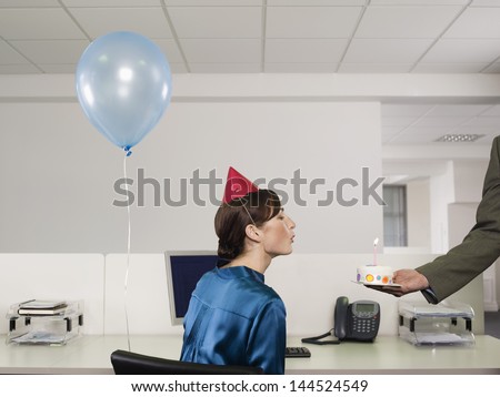 Side view of a woman in party hat blowing birthday candle on cake in office