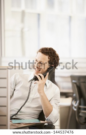 Smiling young male executive sitting in cubicle talking on phone