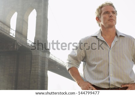 Serious man standing with hands on hip against Brooklyn bridge