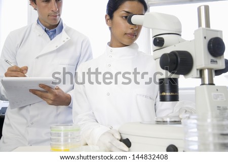 Female scientist adjusting microscope with male colleague noting down in laboratory