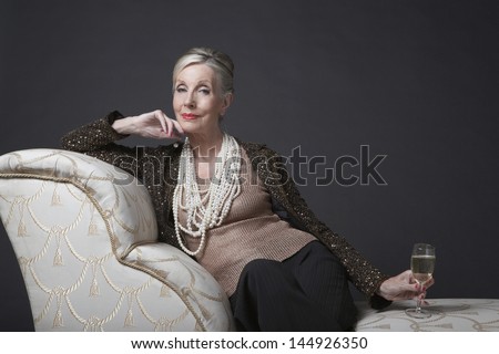 Portrait of an elegant senior woman sitting on chaise lounge with champagne against black background