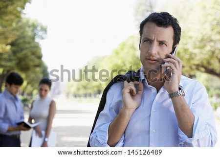 Handsome young businessman looking away while using mobile phone on road with people in background