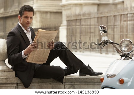 Young businessman sitting by scooter reading newspaper