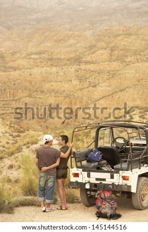 Rear view of affectionate couple looking at desert from cliff standing by four-wheel-drive car