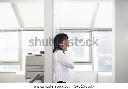 Side view of happy young businesswoman using cellphone leaning on partition in office