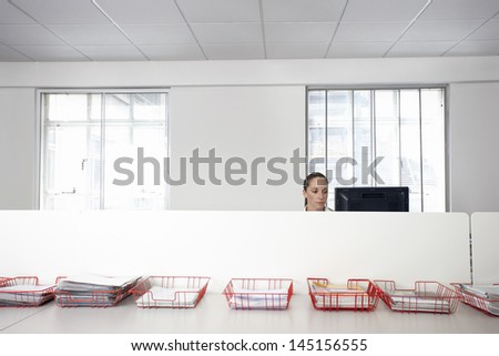 Businesswoman using computer in office cubicle behind trays with documents