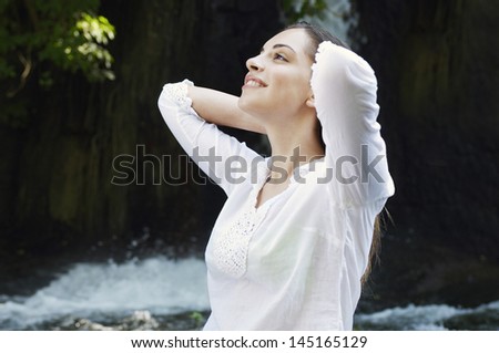 Young woman with hands behind head smiling while standing against waterfall