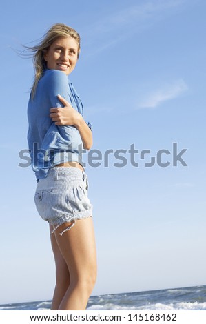 Low angle view of young woman with arms crossed standing against sky at beach