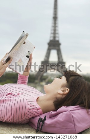 Side view of a young woman lying on balcony and reading book in front of Eiffel Tower