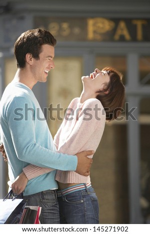 Side view of a laughing couple with shopping bags embracing in front of shop