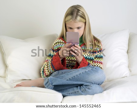 Young girl sitting barefoot on sofa and playing handheld video game