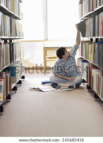 Male student sitting on floor and reaching for book in library