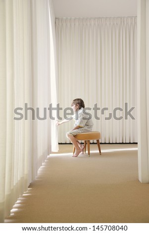 Side view of boy sitting on stool in room looking out window