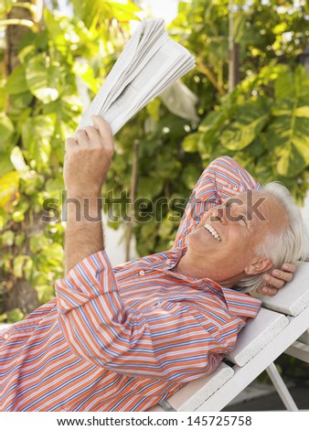 Side view of a smiling mature man reclining on lounge chair and reading newspaper