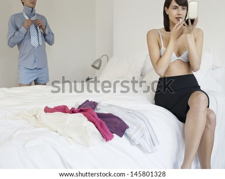 Semi dressed woman applying lipstick on bed with man getting dressed in background