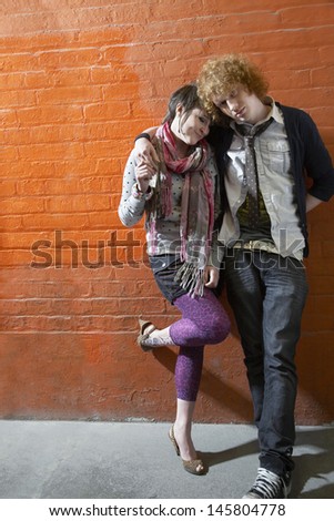 Full length of young loving couple with arm around leaning on orange brick wall
