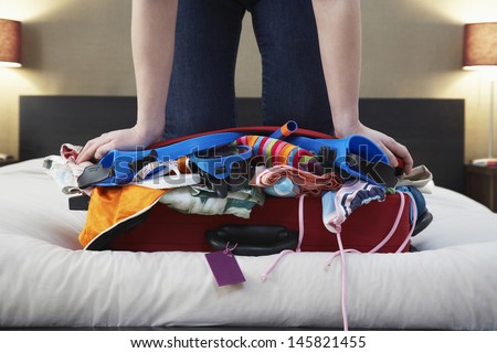 Low section of woman kneeling on overstuffed suitcase in bed