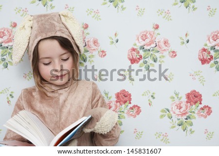 Young girl in bunny costume reading book against wallpaper with floral pattern