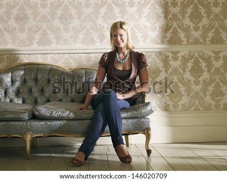 Full length portrait of a young woman sitting on sofa against wallpaper