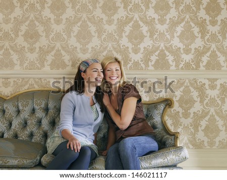 Two cheerful young women sitting on sofa against wallpaper
