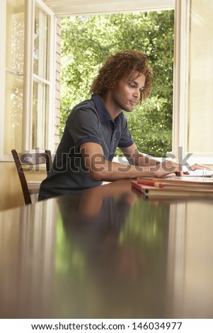 Side view of a young man reading at table by window in living room