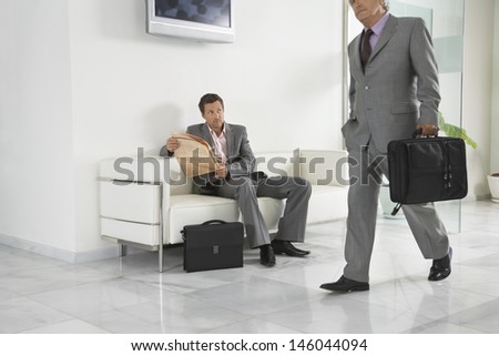 Smart senior businessman walking past a man with newspaper in office hallway