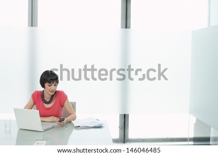 Beautiful casual young woman sitting with laptop and using cellphone in modern cubicle at office
