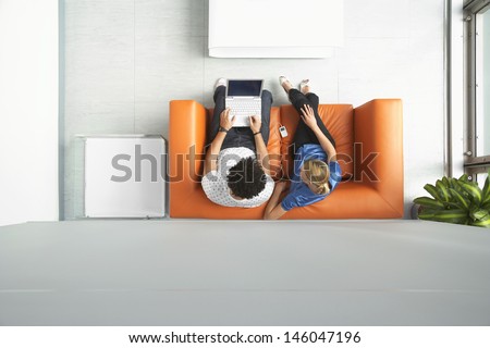 Full length top view of a man and woman using laptop on orange sofa in reception room at office