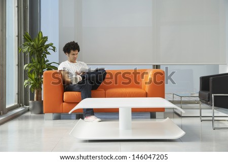 Young man reading newspaper on orange sofa in the reception room at office