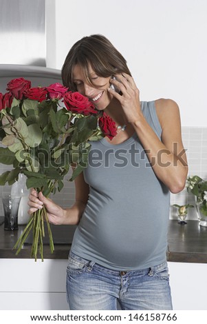 Happy pregnant woman using mobile phone while holding roses in kitchen