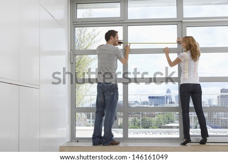 Full length rear view of young couple measuring window in modern apartment