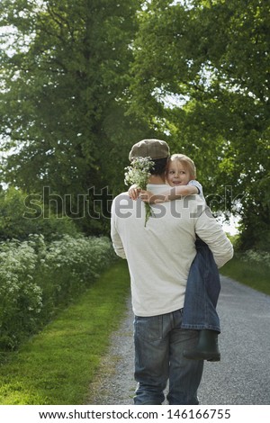 Rear view of a father carrying daughter with flowers on country lane
