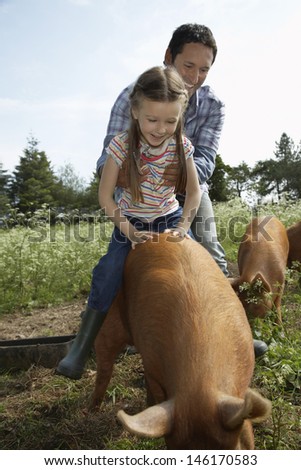 Smiling father helping daughter to ride pig in sty against the sky