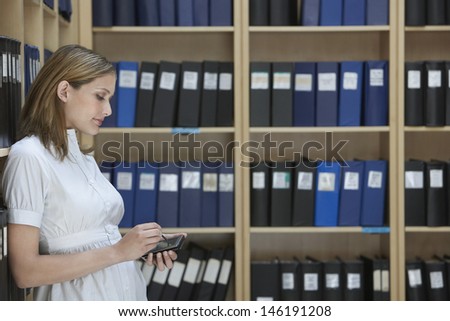 Side view of a young female office worker using palm top in file storage room