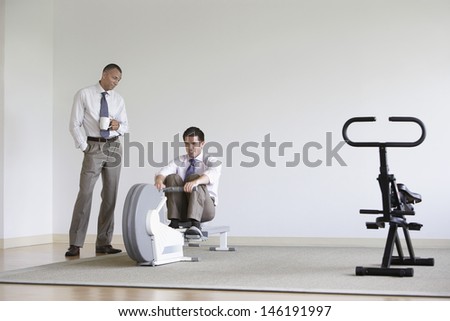 Businessman using rowing machine with colleague standing by