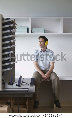 Full length of a man with ear protectors sitting by photocopier in office