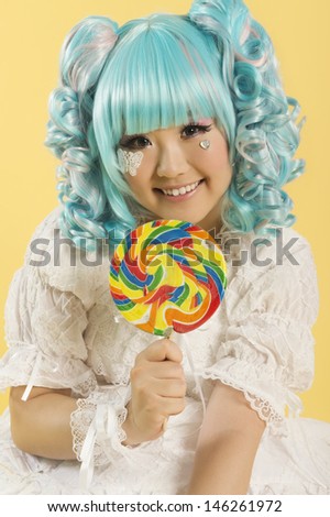 Portrait of smiling young woman dressed as a doll holding lollipop over yellow background