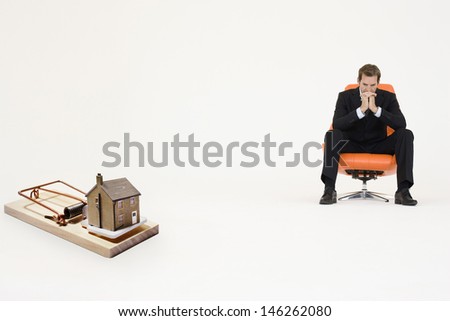 Model home on mouse trap with worried businessman sitting on chair representing increasing real estate rates