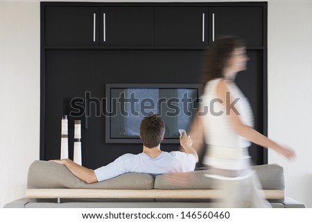 Blurred motion of woman passing by man using remote control while watching TV at home