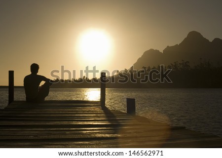 Rear view of man sitting on dock by lake enjoying sunset