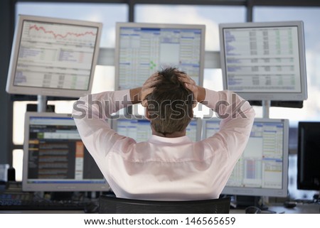 Rear view of stock trader with hands on head looking at multiple computer screens