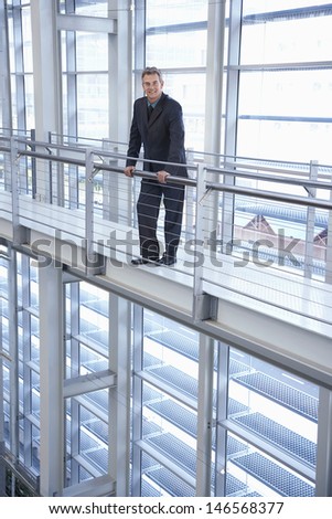 Full length portrait of happy middle aged businessman standing by railing in modern office