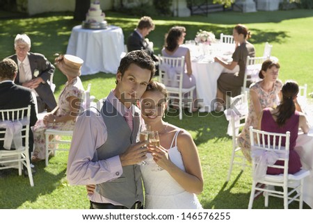 Portrait of young newlywed couple toasting champagne while wedding guests sitting in garden