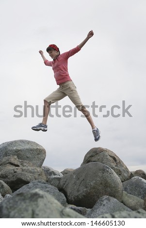 Full length low angle view of a young woman jumping with arms raised over rocks
