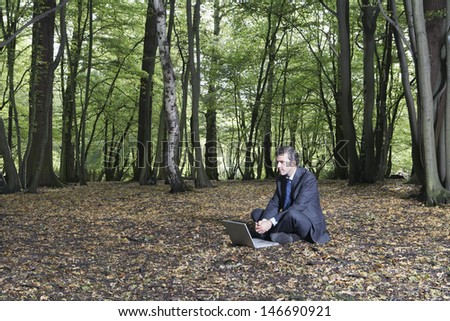 Businessman looking away while sitting with laptop on land in forest